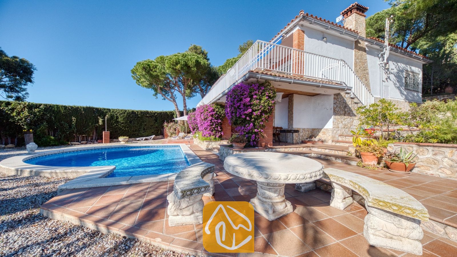 Vakantievilla's Costa Brava Spanje - Villa Gabriella - Privézwembad bij Villa Gabriella, voorzien van een gebeeldhouwde stenen tafel en bijpassende banken op een terras van terracotta. Paarse bougainville klimt tegen de witte muren, onder een wit balkonhek.