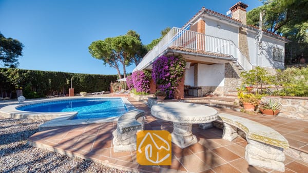 Vakantievilla's Costa Brava Spanje - Villa Gabriella - Privézwembad bij Villa Gabriella, voorzien van een gebeeldhouwde stenen tafel en bijpassende banken op een terras van terracotta. Paarse bougainville klimt tegen de witte muren, onder een wit balkonhek.