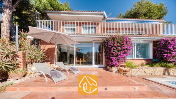 Vakantievilla's Costa Brava Spanje - Villa Gabriella - Zonneterras bij Villa Gabriella met grijze ligstoelen onder een grote beige parasol, terracotta tegels en levendige paarse bougainvillea tegen een gevel van rode baksteen.