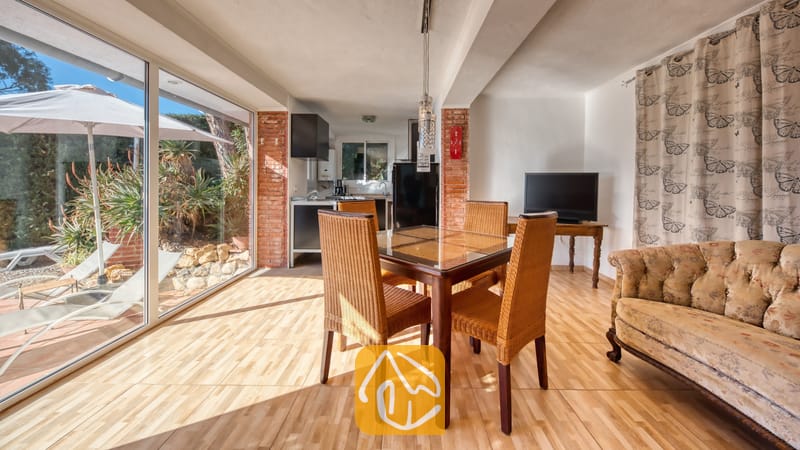 Vakantievilla's Costa Brava Spanje - Villa Gabriella - Eethoek met een glazen tafel en rieten stoelen in Villa Gabriella, voorzien van een bank met patroon en glazen deuren die toegang bieden tot een terras.