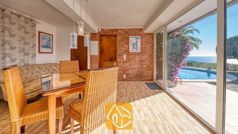 Vakantievilla's Costa Brava Spanje - Villa Gabriella - Eethoek in Villa Gabriella met rieten stoelen en een glazen tafel. Schuifpuien bieden toegang tot een terras met zwembad, een palmboom en uitzicht op zee.