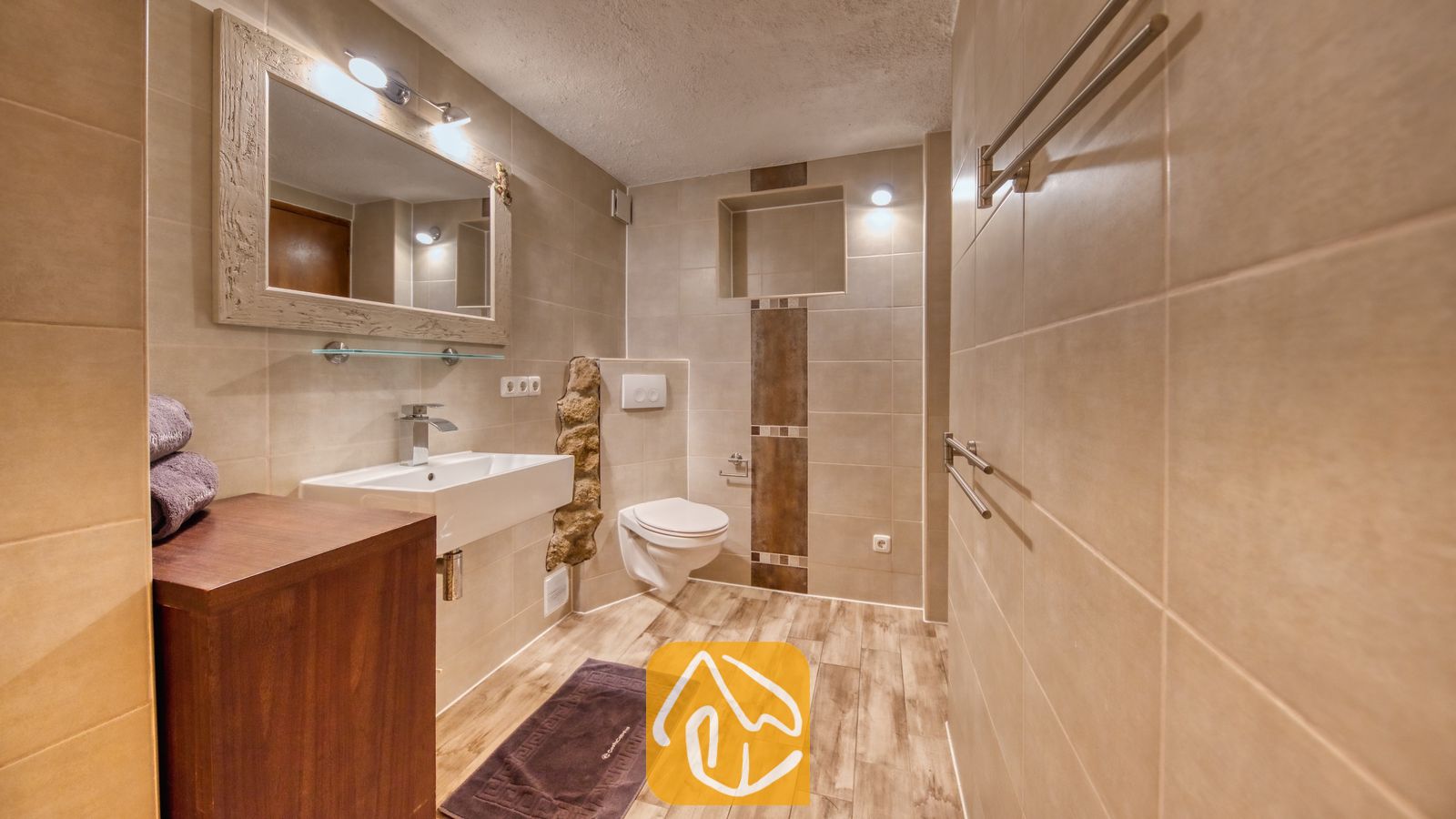 Vakantievilla's Costa Brava Spanje - Villa Gabriella - De badkamer van Villa Gabriella is voorzien van een witte wastafel, een hangend toilet en vloertegels met houtlook. Boven het wastafelmeubel hangt een grote spiegel met omlijsting en geïntegreerde verlichting.