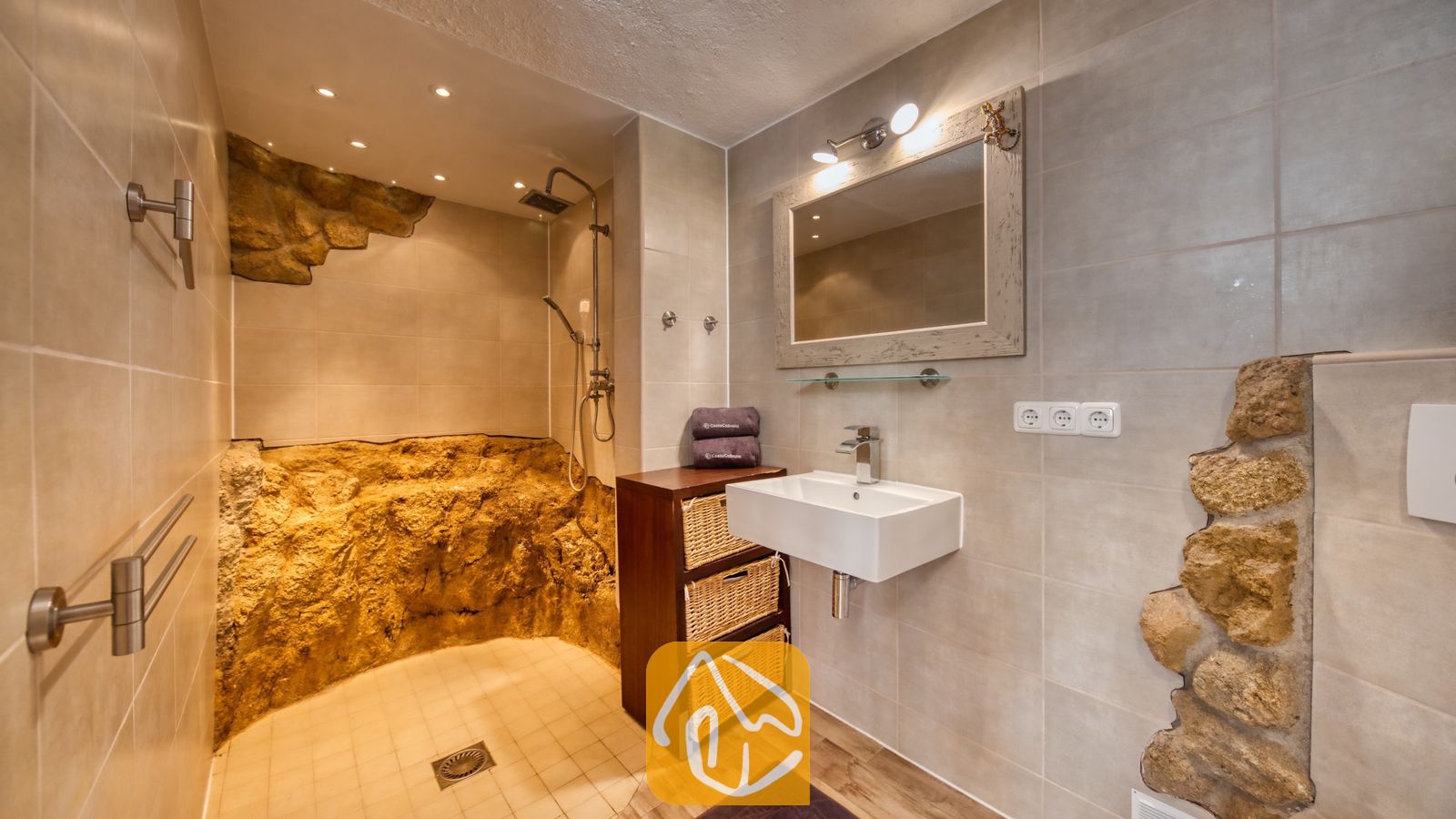 Vakantievilla's Costa Brava Spanje - Villa Gabriella - Badkamer met inloopdouche voorzien van natuurstenen wanden, een witte rechthoekige wastafel en een houten wastafelmeubel met rieten opbergmanden.