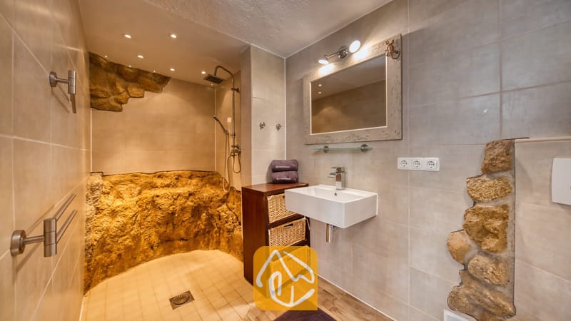 Villas de vacances Costa Brava Espagne - Villa Gabriella - Salle de bain équipée d'une douche à l'italienne avec murs en pierre naturelle, d'un lavabo rectangulaire blanc et d'un meuble sous-vasque en bois agrémenté de paniers de rangement en osier.