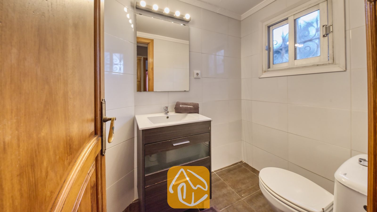 Vakantievilla's Costa Brava Spanje - Villa Abigail - Badkamer met een wastafelmeubel van donker hout, wit getegelde muren, een vierkante spiegel met vijf lampen en een wit toilet.