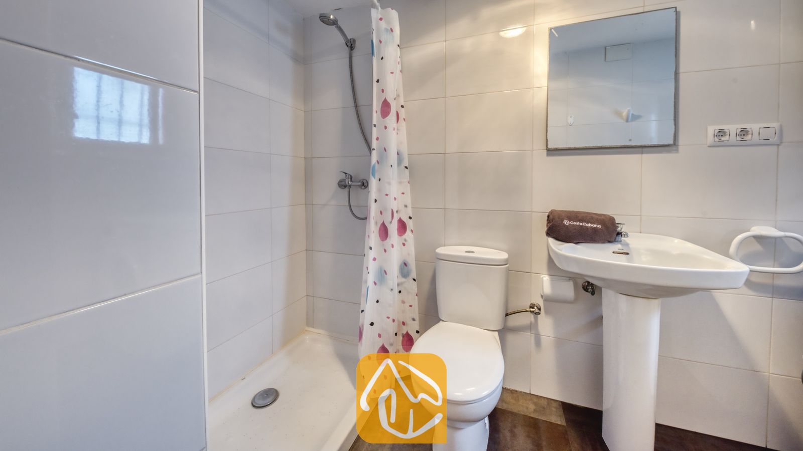 Vakantievilla's Costa Brava Spanje - Villa Abigail - Wit getegelde badkamer met een inloopdouche voorzien van een gordijn met patroon, een toilet en een wastafel op zuil met een opgevouwen bruine handdoek en een wandspiegel.