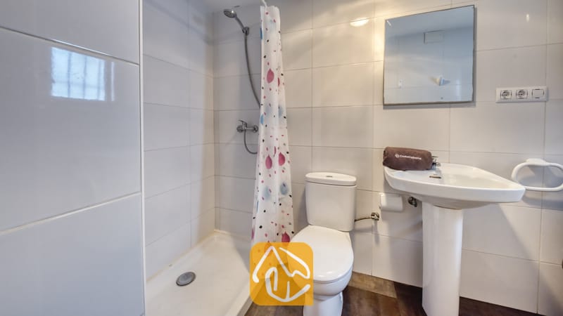 Holiday villas Costa Brava Spain - Villa Abigail - White tiled bathroom featuring a shower stall with a patterned curtain, a toilet, and a pedestal sink with a folded brown towel and wall mirror.