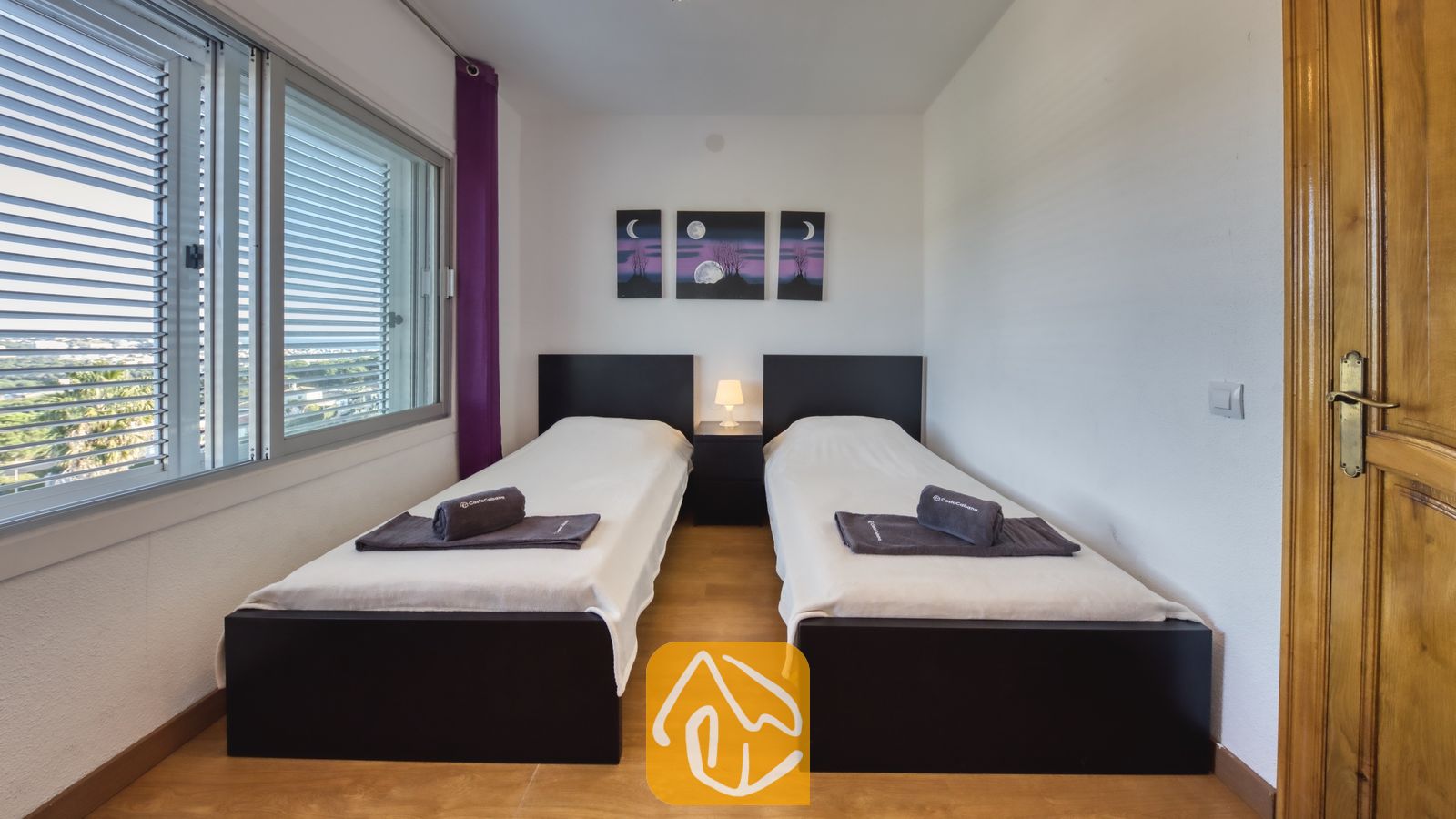 Vakantievilla's Costa Brava Spanje - Villa Abigail - Tweepersoonskamer met twee eenpersoonsbedden met donkerbruine frames, witte beddengoed, opgevouwen grijze handdoeken en een centraal nachtkastje met een klein lampje onder drie kunstpanelen met een maanmotief.