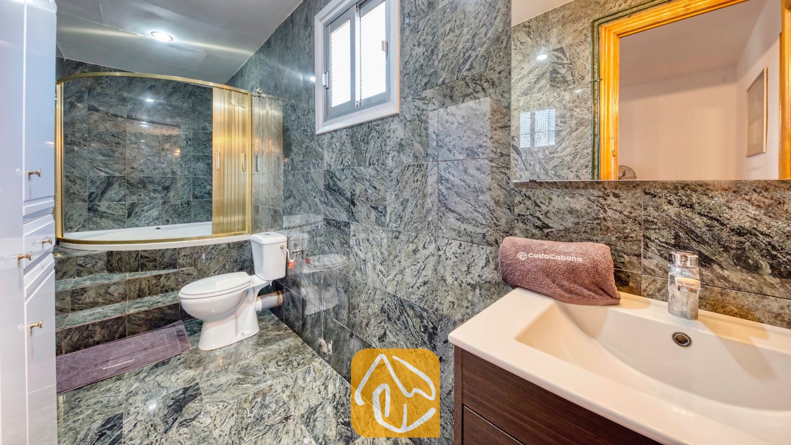 Vakantievilla's Costa Brava Spanje - Villa Abigail - Badkamer voorzien van groen gemarmerde tegelwanden, een wit toilet en een ligbad met een goudomrand glazen scherm.