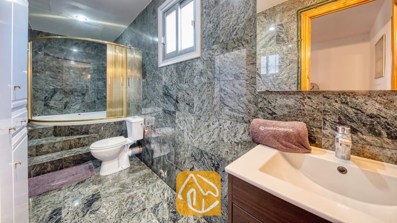 Holiday villas Costa Brava Spain - Villa Abigail - Bathroom featuring green marble-patterned tiled walls, a white toilet, and a bathtub with a gold-framed glass enclosure.
