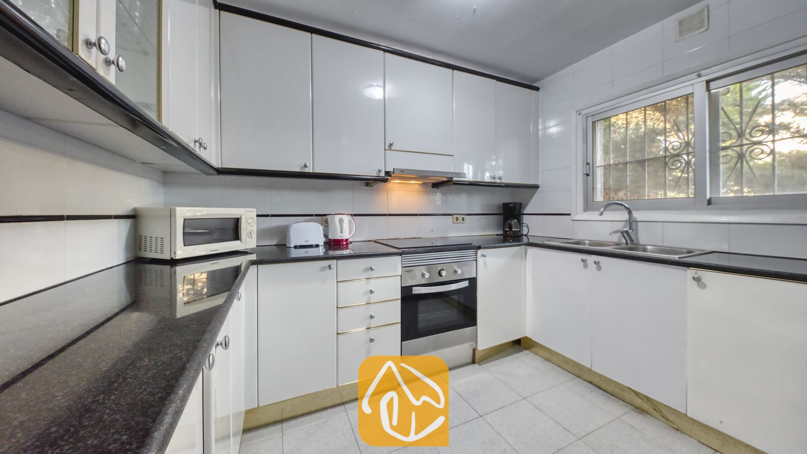 Vakantievilla's Costa Brava Spanje - Villa Abigail - Keuken met hoogglans witte kastjes, zwarte granieten aanrechtbladen, een roestvrijstalen oven en een raam met dievenklauwen.