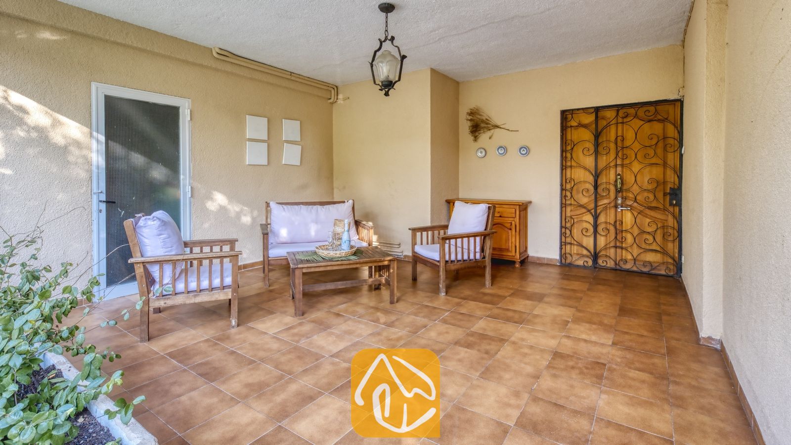 Vakantievilla's Costa Brava Spanje - Villa Abigail - Overdekt terras bij Villa Abigail met houten fauteuils met witte kussens, een salontafel en een decoratieve smeedijzeren deur.