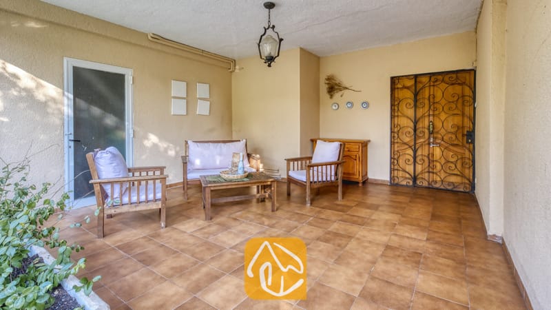 Vakantievilla's Costa Brava Spanje - Villa Abigail - Overdekt terras bij Villa Abigail met houten fauteuils met witte kussens, een salontafel en een decoratieve smeedijzeren deur.