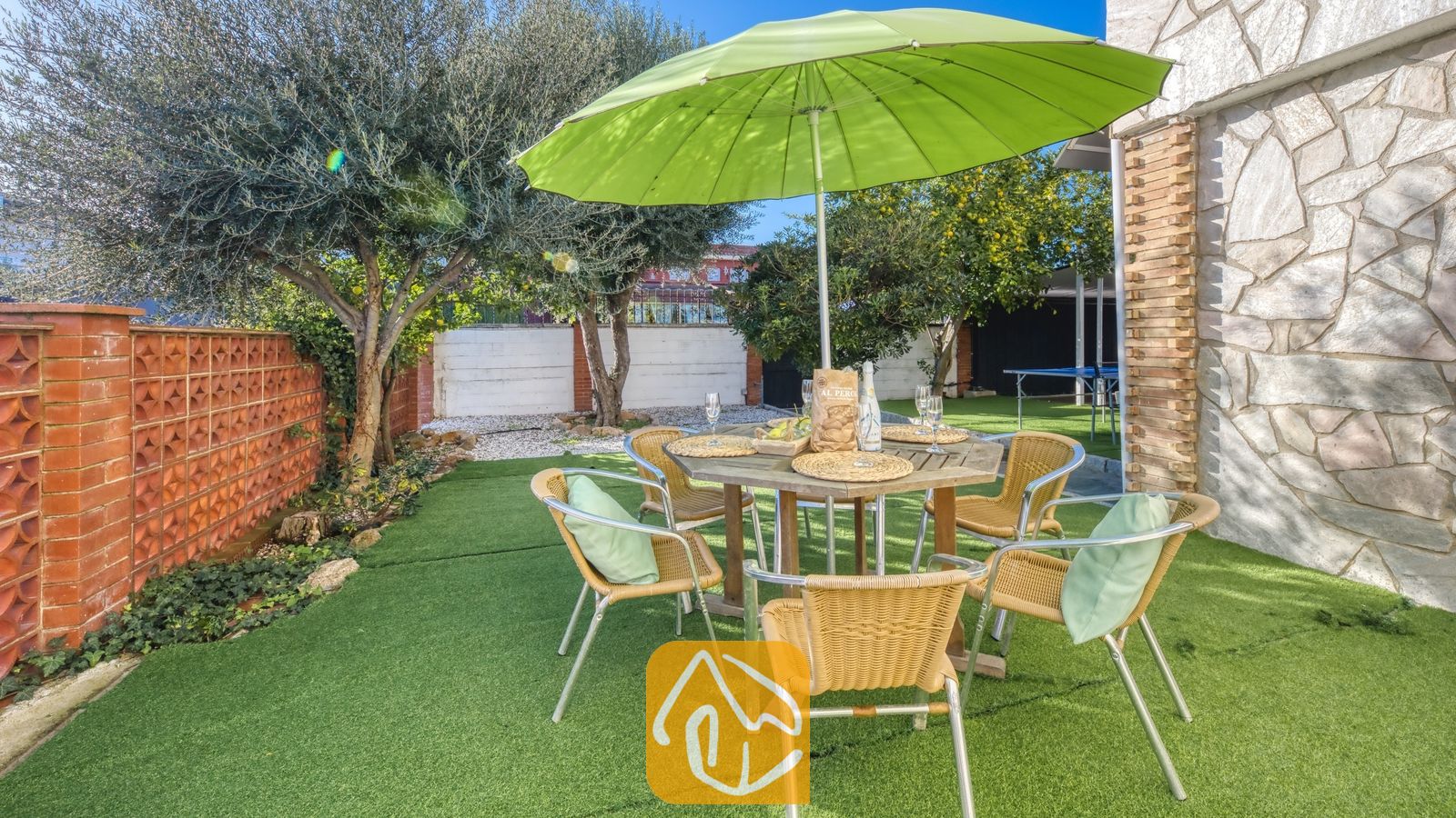 Vakantievilla's Costa Brava Spanje - Villa Abigail - Eethoek in de buitenlucht bij Villa Abigail, voorzien van een houten tafel, geweven stoelen met groene kussens en een grote groene parasol op een kunstgrasgazon.