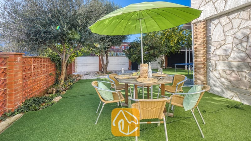 Vakantievilla's Costa Brava Spanje - Villa Abigail - Eethoek in de buitenlucht bij Villa Abigail, voorzien van een houten tafel, geweven stoelen met groene kussens en een grote groene parasol op een kunstgrasgazon.
