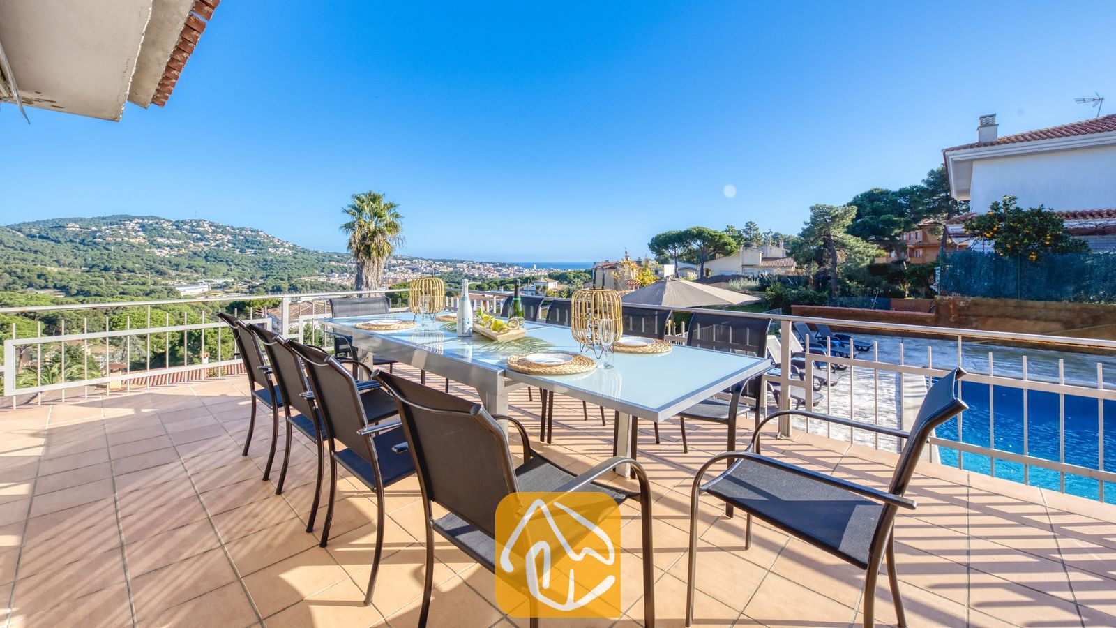 Vakantievilla's Costa Brava Spanje - Villa Abigail - Zonnig terras bij Villa Abigail met een grote glazen eettafel omringd door zwarte gaasstoelen, met uitzicht op verre heuvels en een zwembad.