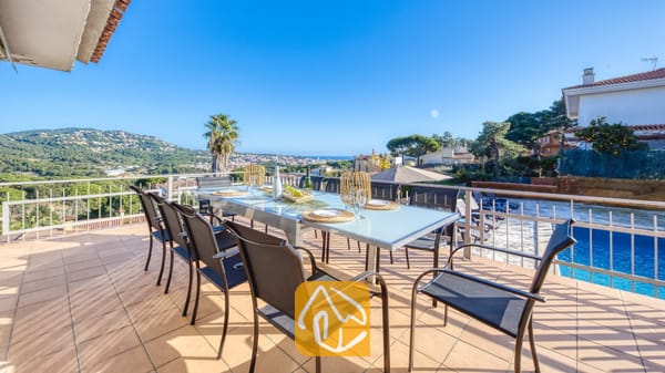 Vakantievilla's Costa Brava Spanje - Villa Abigail - Zonnig terras bij Villa Abigail met een grote glazen eettafel omringd door zwarte gaasstoelen, met uitzicht op verre heuvels en een zwembad.