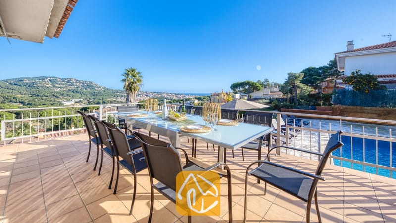 Vakantievilla's Costa Brava Spanje - Villa Abigail - Zonnig terras bij Villa Abigail met een grote glazen eettafel omringd door zwarte gaasstoelen, met uitzicht op verre heuvels en een zwembad.