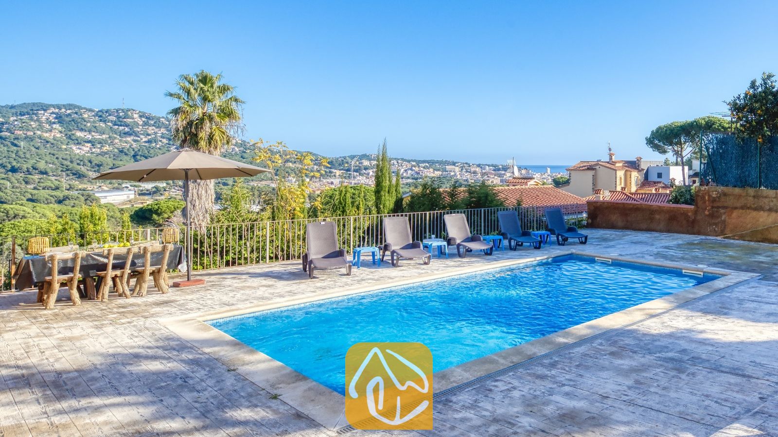 Vakantievilla's Costa Brava Spanje - Villa Abigail - Privézwembad en terras bij Villa Abigail met helderblauw water, rijen zwarte ligbedden, een eettafel voor buiten en een uitzicht in de verte op de bergen onder een strakke, heldere hemel.