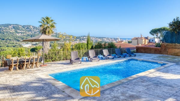 Vakantievilla's Costa Brava Spanje - Villa Abigail - Privézwembad en terras bij Villa Abigail met helderblauw water, rijen zwarte ligbedden, een eettafel voor buiten en een uitzicht in de verte op de bergen onder een strakke, heldere hemel.