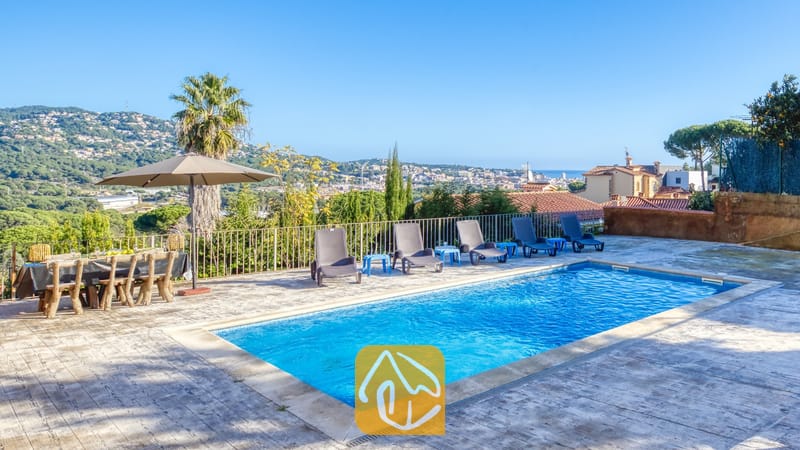 Vakantievilla's Costa Brava Spanje - Villa Abigail - Privézwembad en terras bij Villa Abigail met helderblauw water, rijen zwarte ligbedden, een eettafel voor buiten en een uitzicht in de verte op de bergen onder een strakke, heldere hemel.