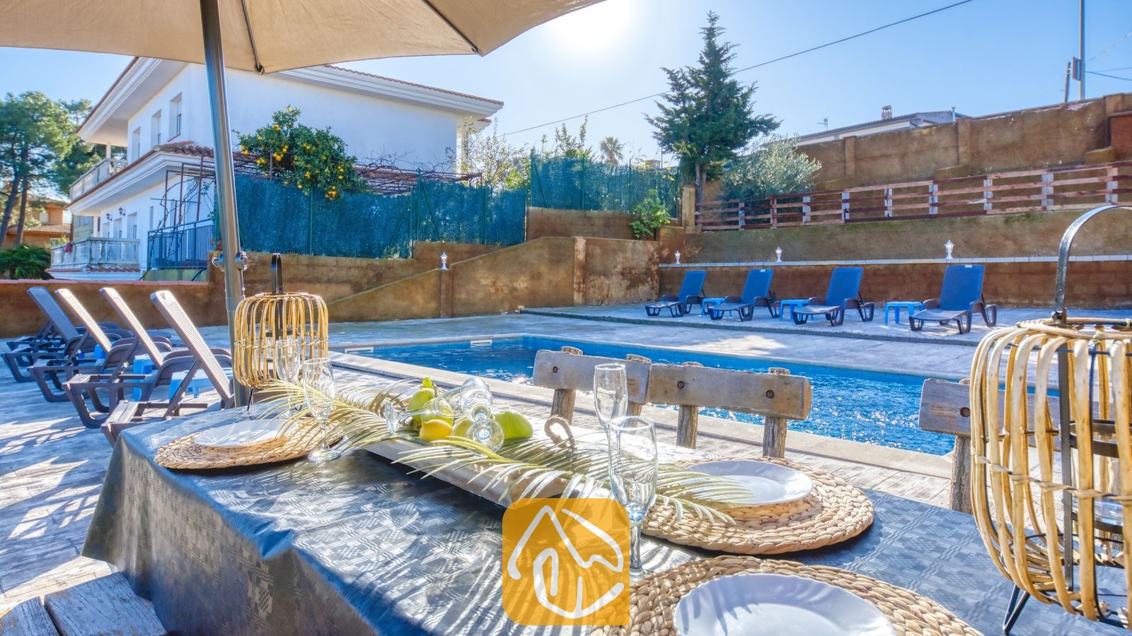 Vakantievilla's Costa Brava Spanje - Villa Abigail - Een eettafel in de buitenlucht bij het zwembad van Villa Abigail, voorzien van rieten placemats, champagneglazen en geweven lantaarns onder een parasol.