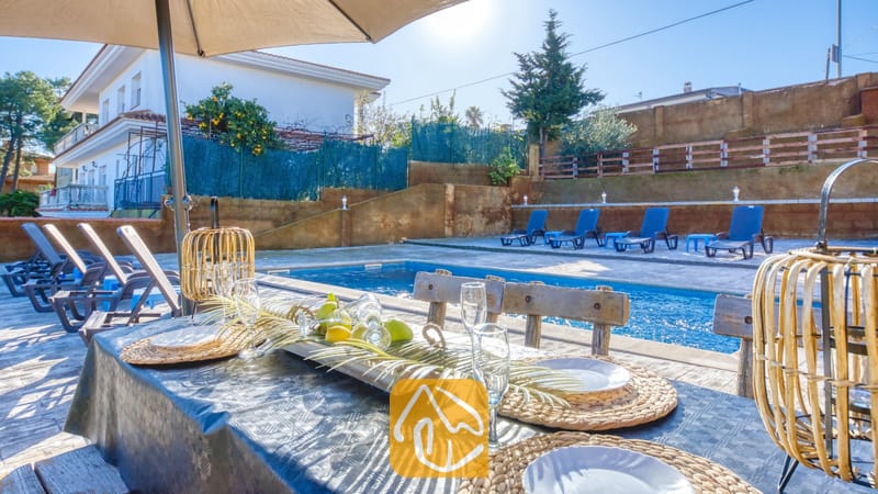 Vakantievilla's Costa Brava Spanje - Villa Abigail - Een eettafel in de buitenlucht bij het zwembad van Villa Abigail, voorzien van rieten placemats, champagneglazen en geweven lantaarns onder een parasol.