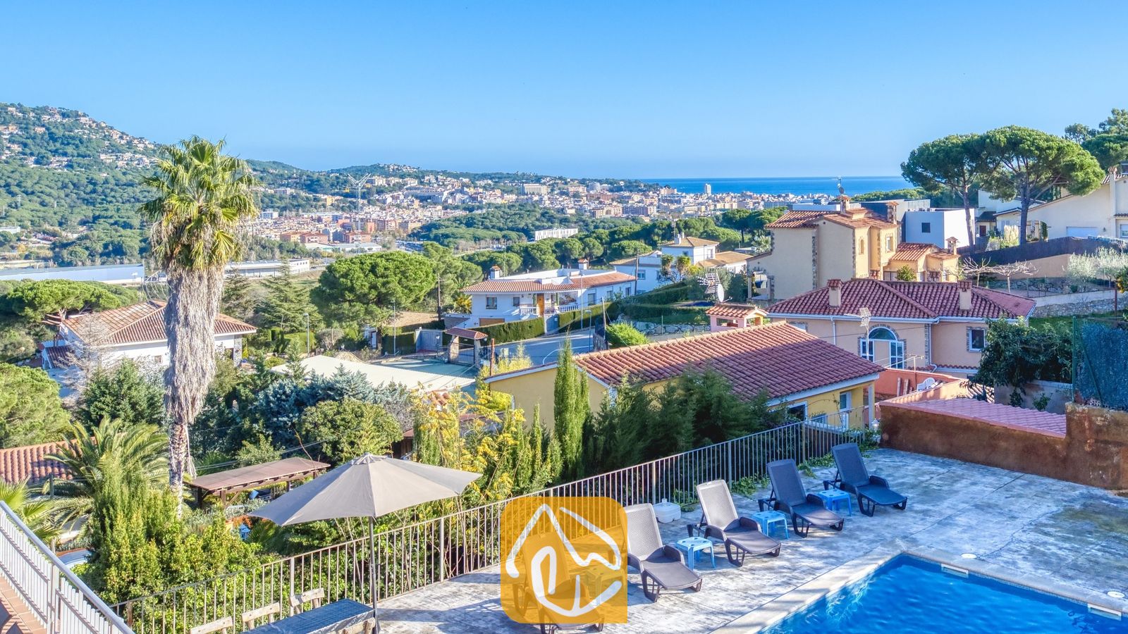 Vakantievilla's Costa Brava Spanje - Villa Abigail - Verheven uitzicht vanaf Villa Abigail over een blauw zwembad, ligbedden en de verre stad en zee onder een heldere hemel.