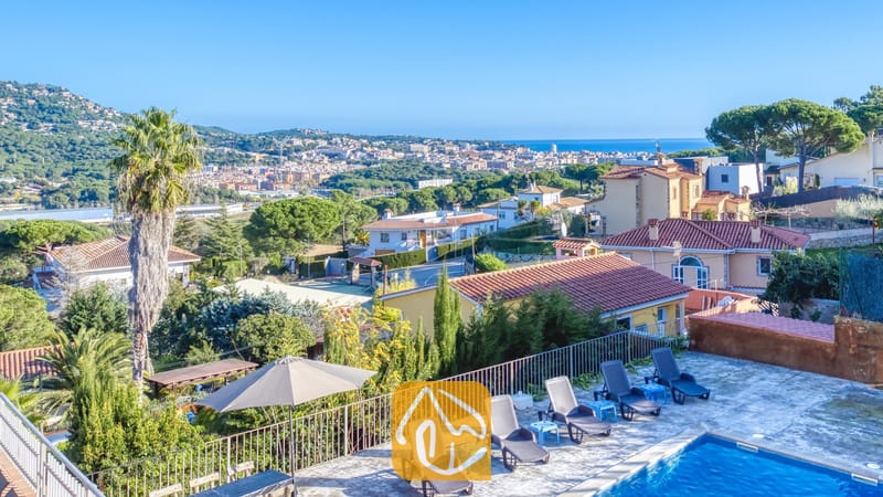 Holiday villas Costa Brava Spain - Villa Abigail - Elevated view from Villa Abigail overlooking a blue swimming pool, sun loungers, and distant town and sea under a clear sky.