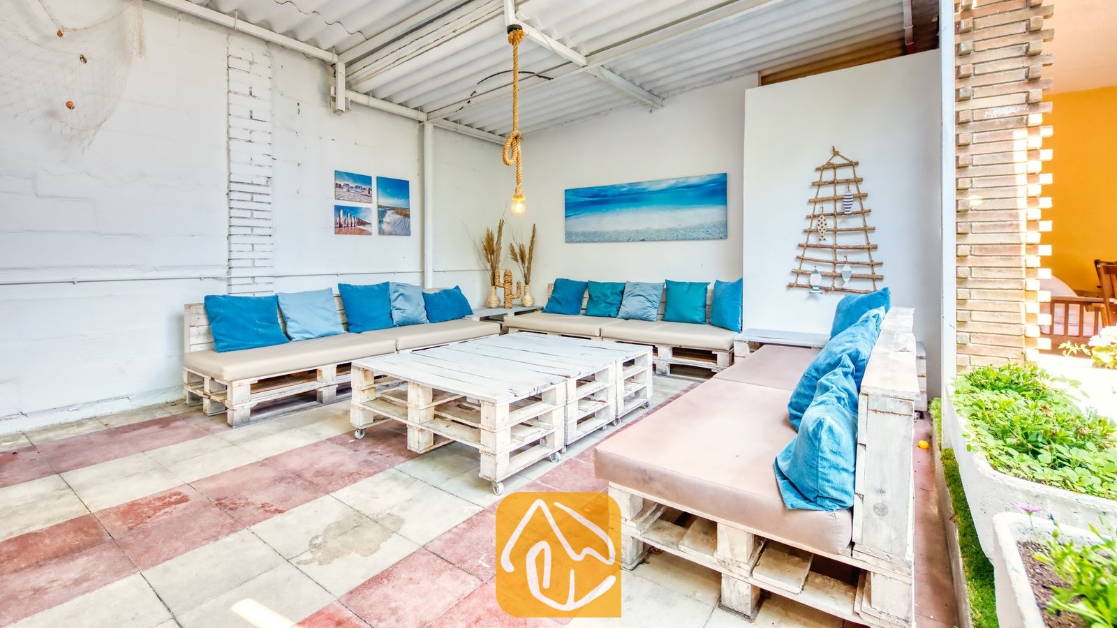 Vakantievilla's Costa Brava Spanje - Villa Abigail - Overdekt buitenterras bij Villa Abigail met handgemaakte witte houten palletbanken voorzien van beige kussens en blauwe sierkussens, lage salontafels van pallets en tegelvloeren.