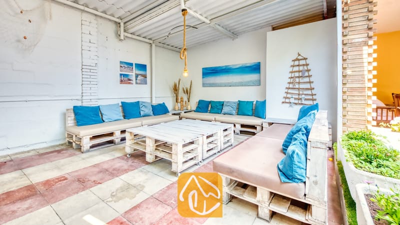 Vakantievilla's Costa Brava Spanje - Villa Abigail - Overdekt buitenterras bij Villa Abigail met handgemaakte witte houten palletbanken voorzien van beige kussens en blauwe sierkussens, lage salontafels van pallets en tegelvloeren.