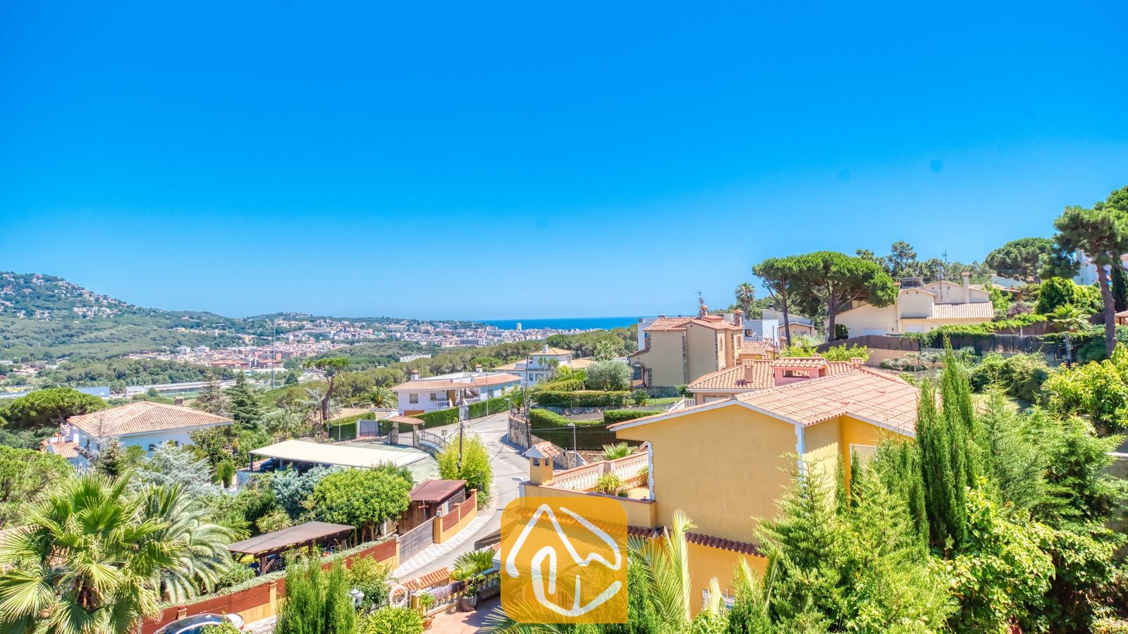 Vakantievilla's Costa Brava Spanje - Villa Abigail - Verheven uitzicht vanuit Villa Abigail over de residentiële heuvels, terracotta daken, weelderige groene bomen en de verre blauwe zee onder een heldere hemel.