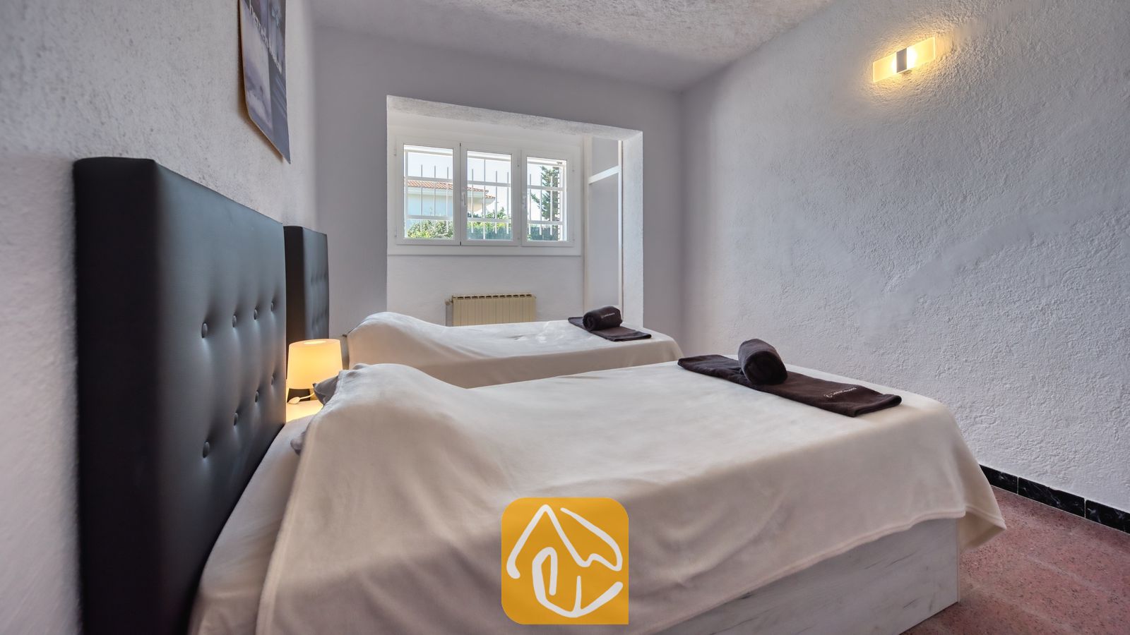 Vakantievilla's Costa Brava Spanje - Villa Abigail - Slaapkamer met twee eenpersoonsbedden voorzien van donkere gecapitonneerde hoofdborden, wit beddengoed en opgerolde donkere handdoeken onder een klein raam.