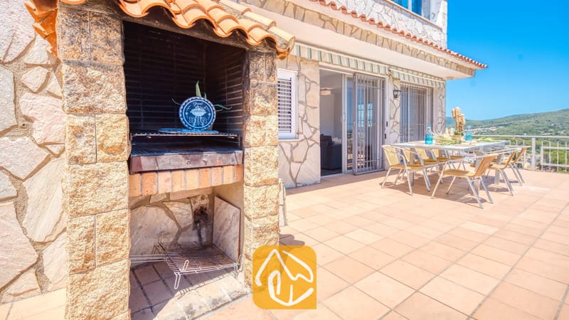 Vakantievilla's Costa Brava Spanje - Villa Abigail - Ingebouwde stenen barbecue op een betegeld terras bij Villa Abigail, voorzien van een eettafel met stoelen buiten en uitzicht op de verre heuvels.