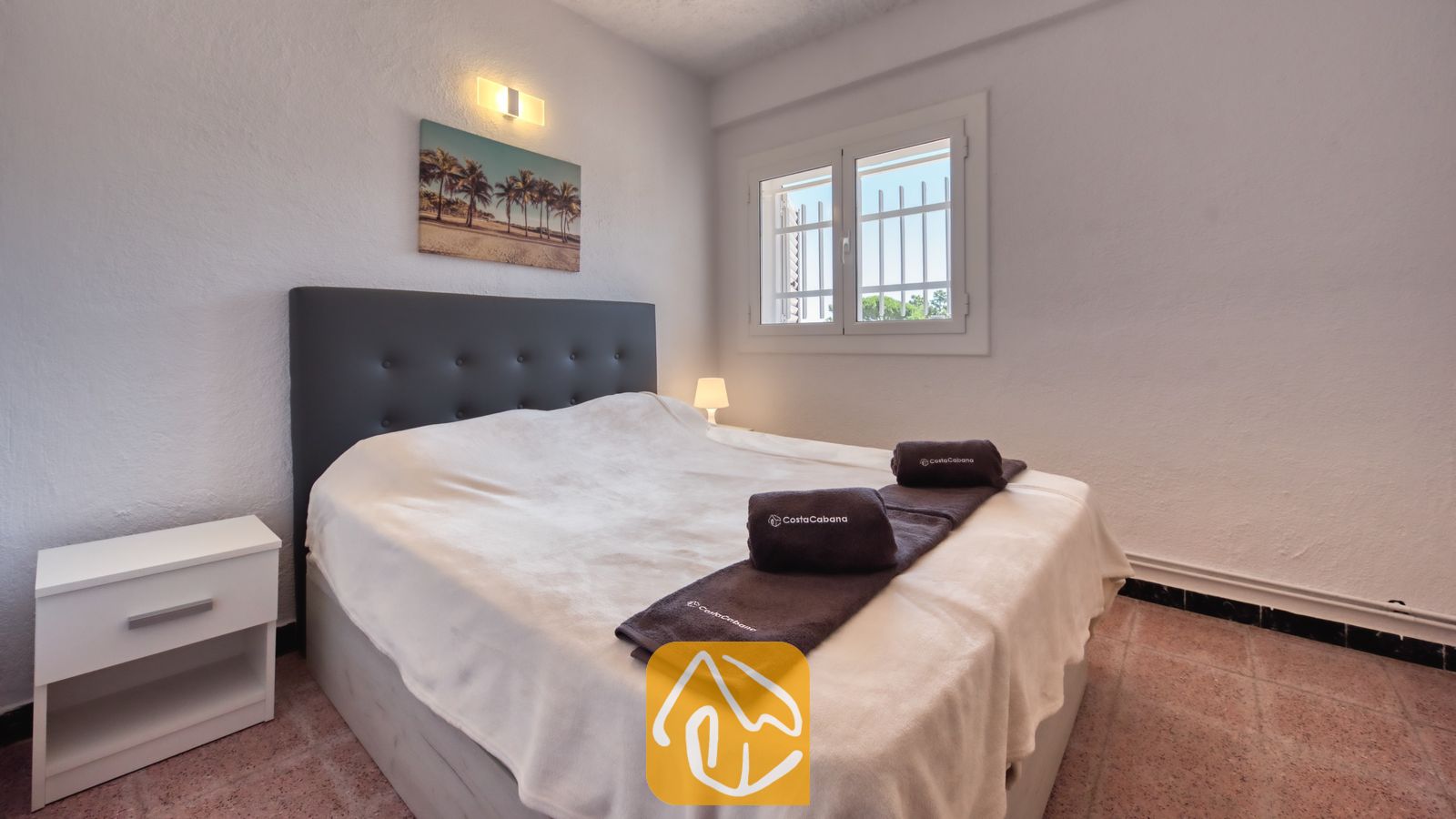 Vakantievilla's Costa Brava Spanje - Villa Abigail - Slaapkamer met een tweepersoonsbed voorzien van een donkergrijs gestoffeerd hoofdbord, opgerolde bruine handdoeken, witte muren en een raam met tralies.