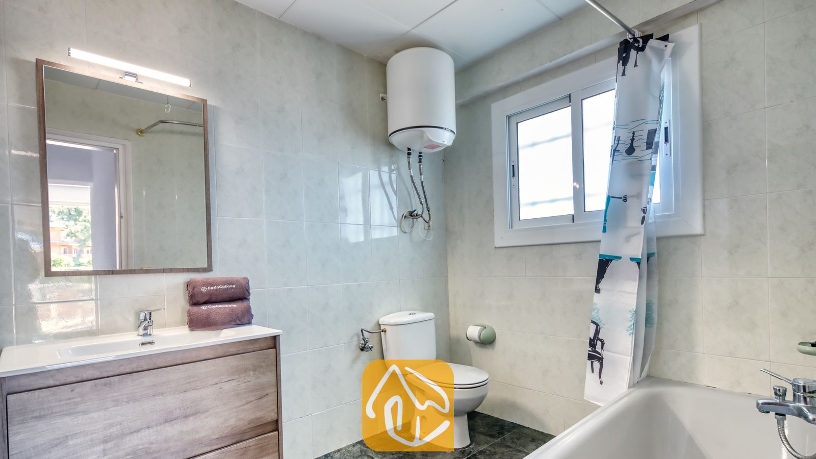 Vakantievilla's Costa Brava Spanje - Villa Abigail - Badkamer met een wit toilet, een houten badkamermeubel met wastafel, en een ligbad met een gedecoreerd douchegordijn onder een raam.