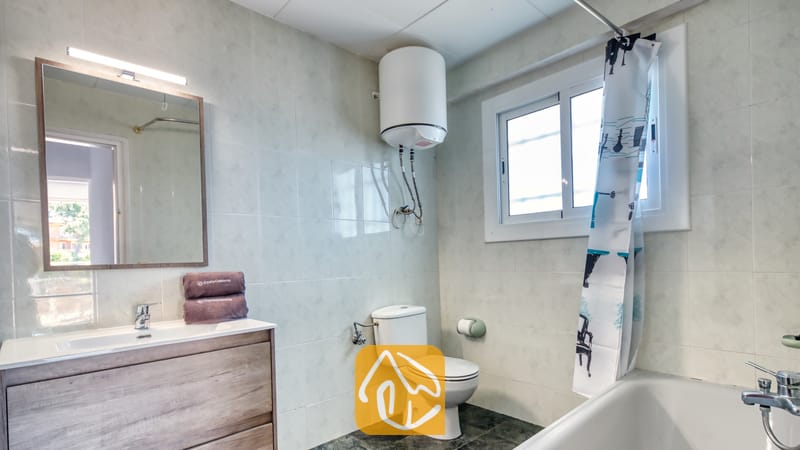 Holiday villas Costa Brava Spain - Villa Abigail - Bathroom with a white toilet, wooden vanity with a washbasin, and a bathtub with a patterned shower curtain under a window.