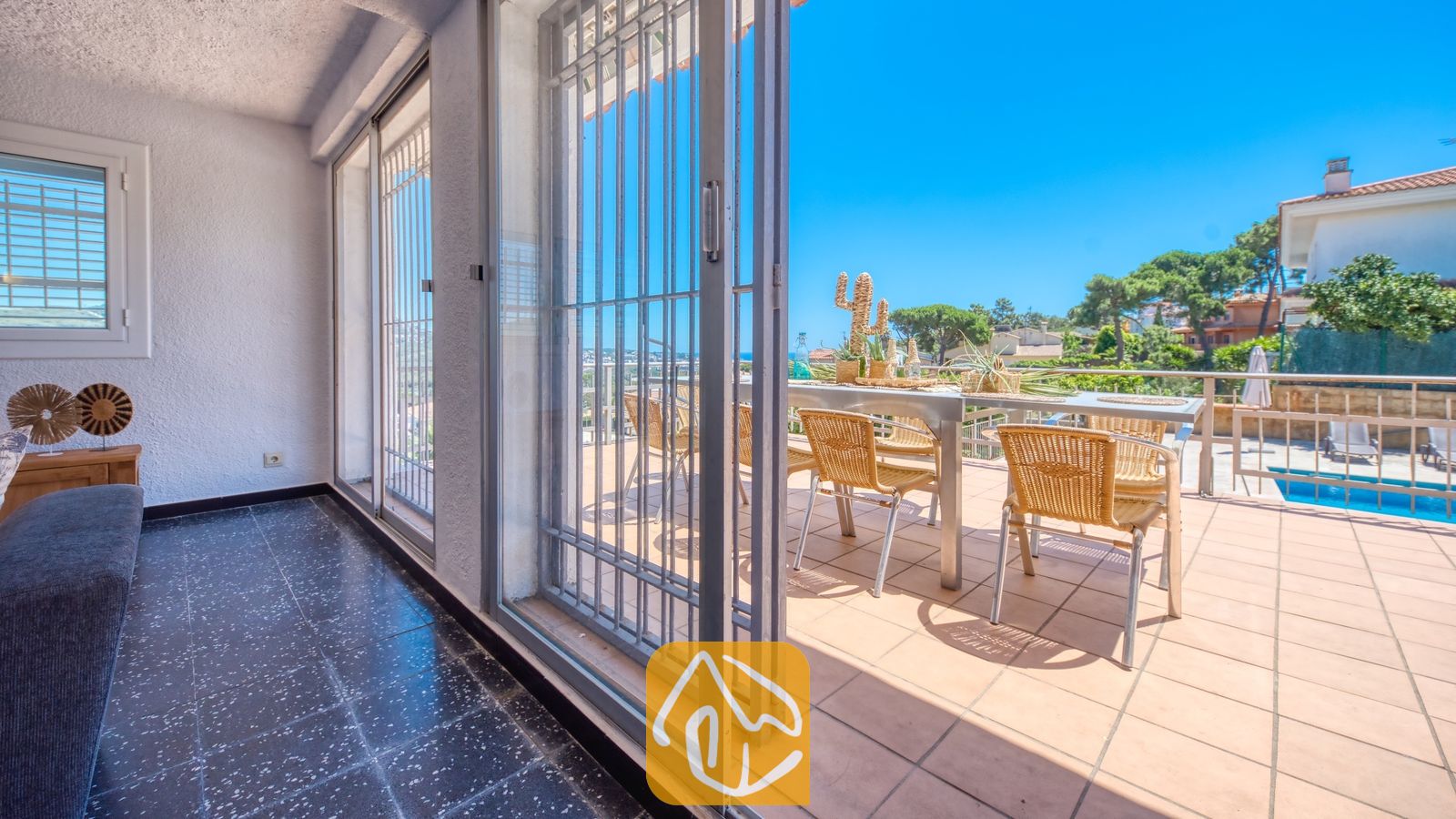 Vakantievilla's Costa Brava Spanje - Villa Abigail - Overdekt terras met donkere tegelvloer dat via glazen schuifpuien uitkomt op een buiten-eethoek met rieten stoelen en uitzicht op het zwembad bij Villa Abigail.
