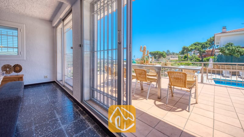 Holiday villas Costa Brava Spain - Villa Abigail - Covered terrace with dark tiled flooring opening via sliding glass doors to an outdoor dining area with wicker chairs and a swimming pool view at Villa Abigail.