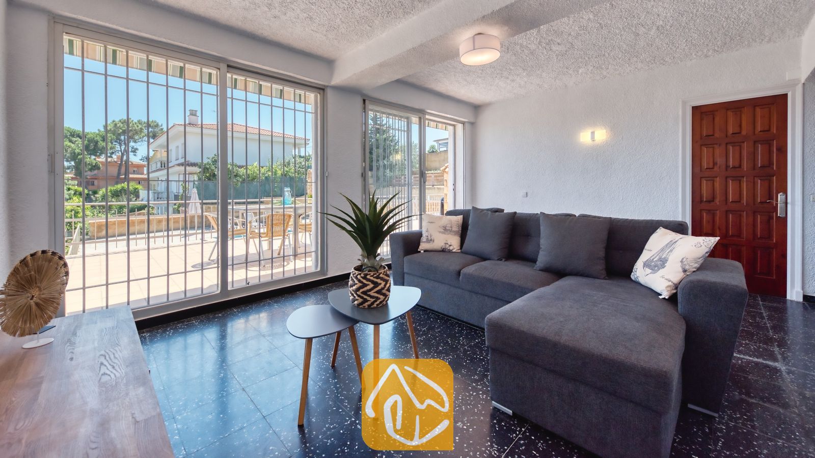 Vakantievilla's Costa Brava Spanje - Villa Abigail - Woonkamer met een grijze hoekbank, donkere tegelvloeren en grote glazen schuifpui die toegang biedt tot een buitenterras.