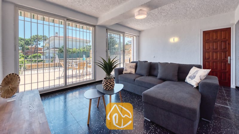 Holiday villas Costa Brava Spain - Villa Abigail - Living room with a grey sectional sofa, dark tiled floors, and large glass sliding doors opening to an outdoor terrace.