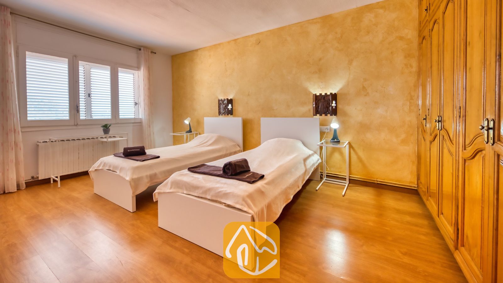 Vakantievilla's Costa Brava Spanje - Villa Abigail - Slaapkamer met twee eenpersoonsbedden met witte frames, opgerolde bruine handdoeken, warme gele, getextureerde muren en een grote houten kledingkast.