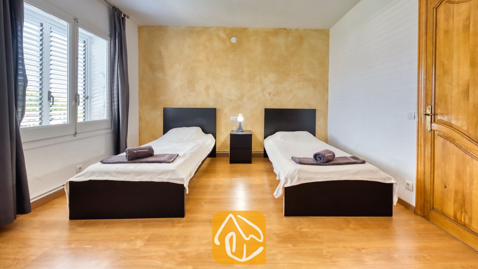Vakantievilla's Costa Brava Spanje - Villa Abigail - Tweepersoonskamer met twee eenpersoonsbedden in Villa Abigail, voorzien van wit beddengoed, donkerbruine hoofdborden en opgevouwen bruine handdoeken, gescheiden door een klein nachtkastje met een lamp.