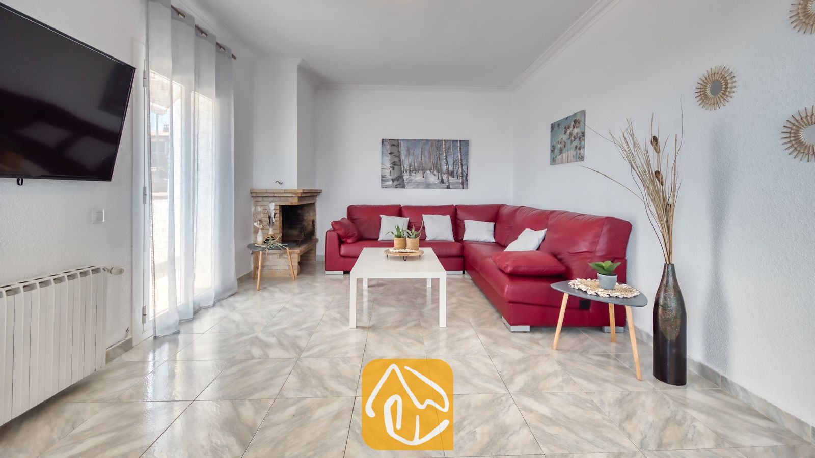 Vakantievilla's Costa Brava Spanje - Villa Abigail - Interieur van de woonkamer in Villa Abigail, voorzien van een grote rode hoekbank, een witte salontafel, tegelvloeren en een stenen open haard.
