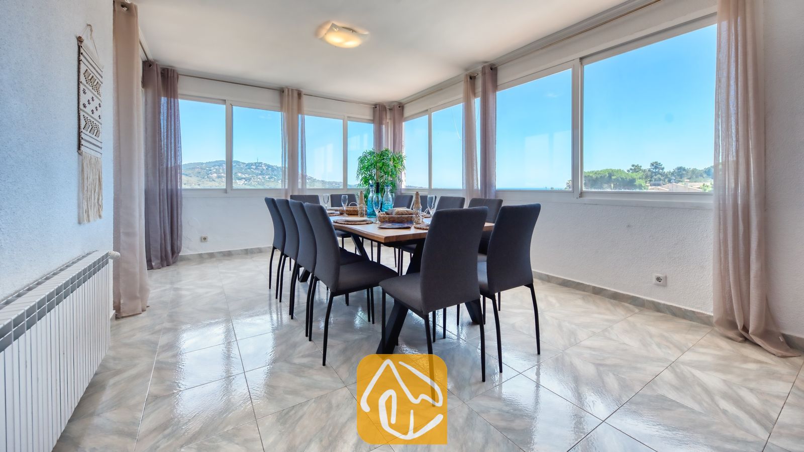 Vakantievilla's Costa Brava Spanje - Villa Abigail - Eetkamer met een lange houten tafel, donkergrijze gestoffeerde stoelen, tegelvloeren en grote ramen met uitzicht naar buiten in Villa Abigail.