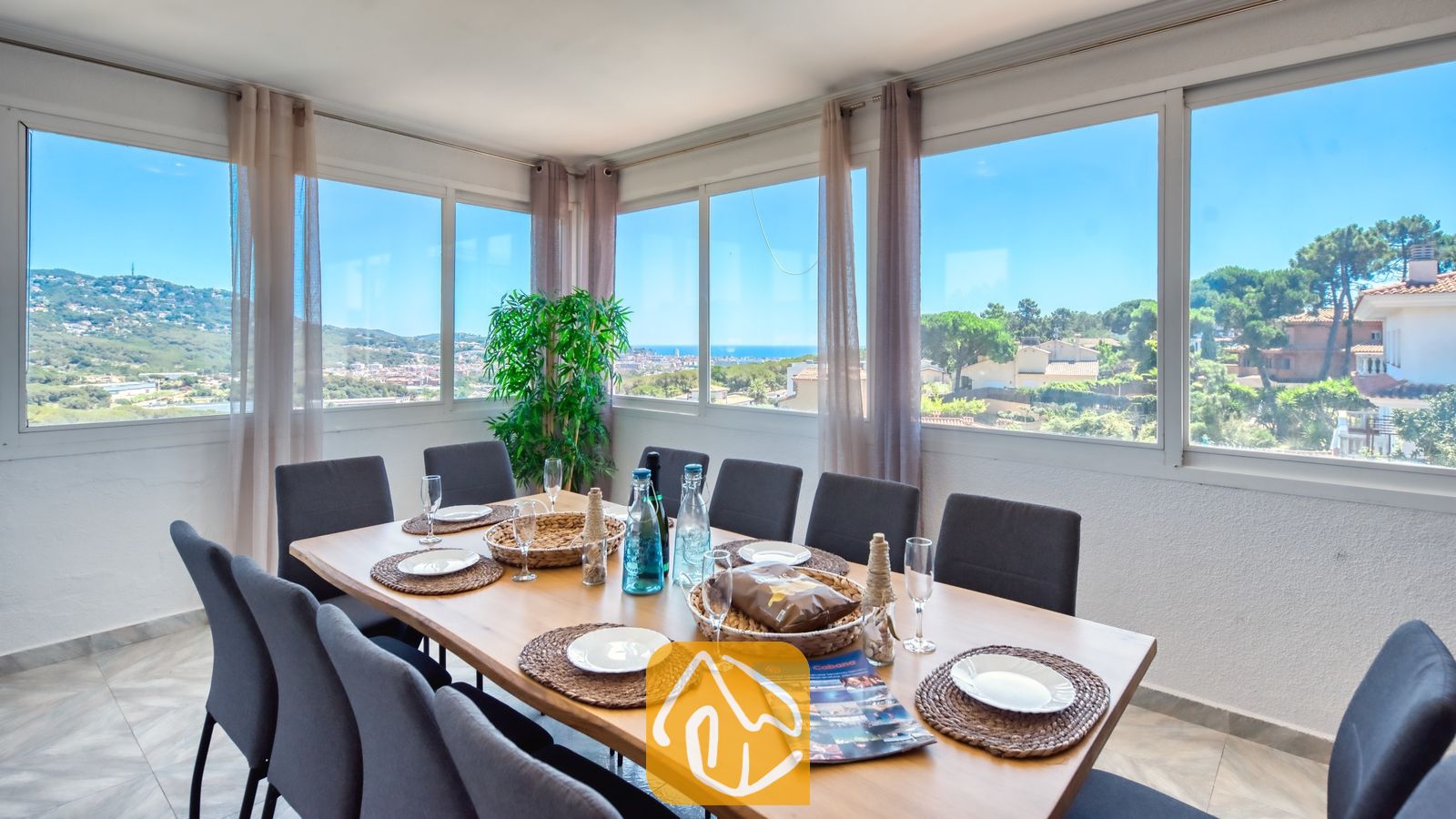 Vakantievilla's Costa Brava Spanje - Villa Abigail - Eethoek met grote ramen en uitzicht op de kust, voorzien van een houten tafel gedekt met borden, geweven placemats en glaswerk in Villa Abigail.