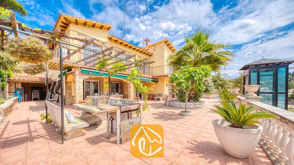Vakantievilla's Costa Brava Spanje - Villa Dolce Vita - Zonnig terras van Villa Dolce Vita met een stenen eettafel, een verrijdbare barbecue, een houten pergola, potten met palmbomen en gele muren tegen een blauwe hemel.