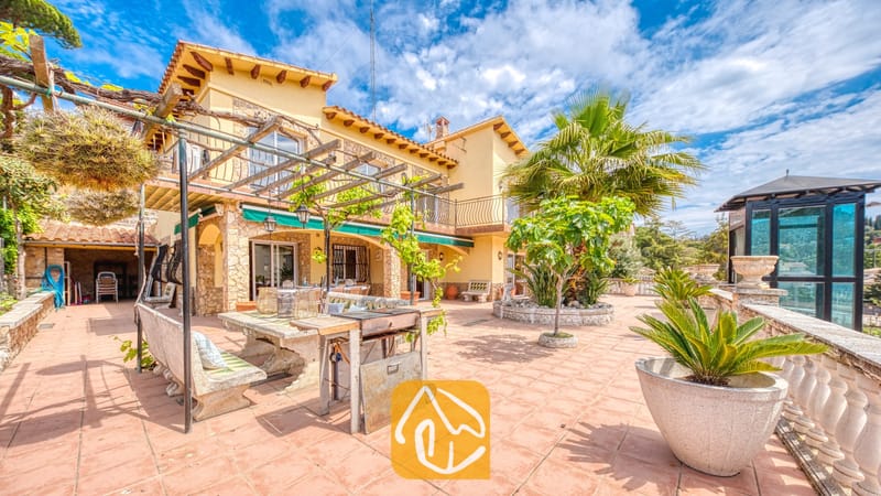 Vakantievilla's Costa Brava Spanje - Villa Dolce Vita - Zonnig terras van Villa Dolce Vita met een stenen eettafel, een verrijdbare barbecue, een houten pergola, potten met palmbomen en gele muren tegen een blauwe hemel.