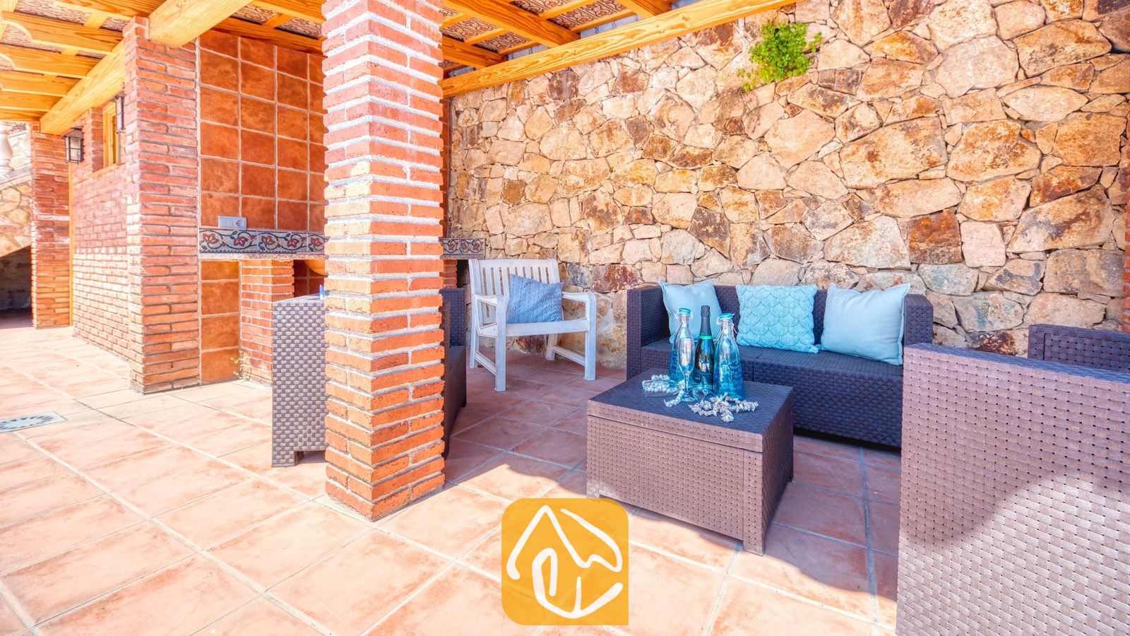 Vakantievilla's Costa Brava Spanje - Villa Dolce Vita - Overdekt buitenterras bij Villa Dolce Vita met bruine rieten zitmeubels, blauwe kussens, een salontafel met glazen flessen en een rustieke stenen muur.