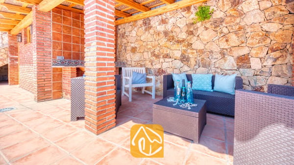 Vakantievilla's Costa Brava Spanje - Villa Dolce Vita - Overdekt buitenterras bij Villa Dolce Vita met bruine rieten zitmeubels, blauwe kussens, een salontafel met glazen flessen en een rustieke stenen muur.